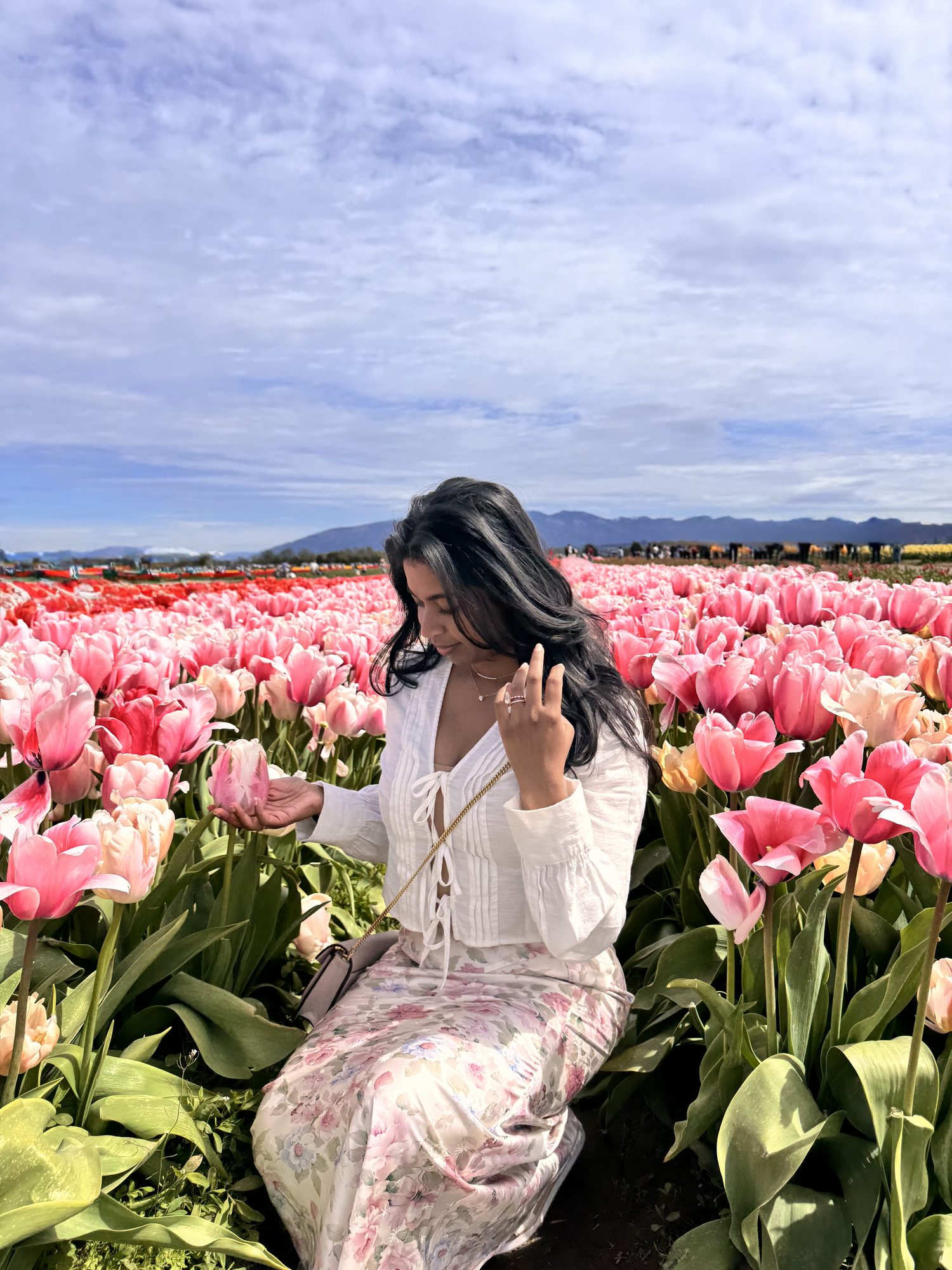 Bhagmattie sitting outdoors in a field of pink tulips, wearing a white top, in soft natural light